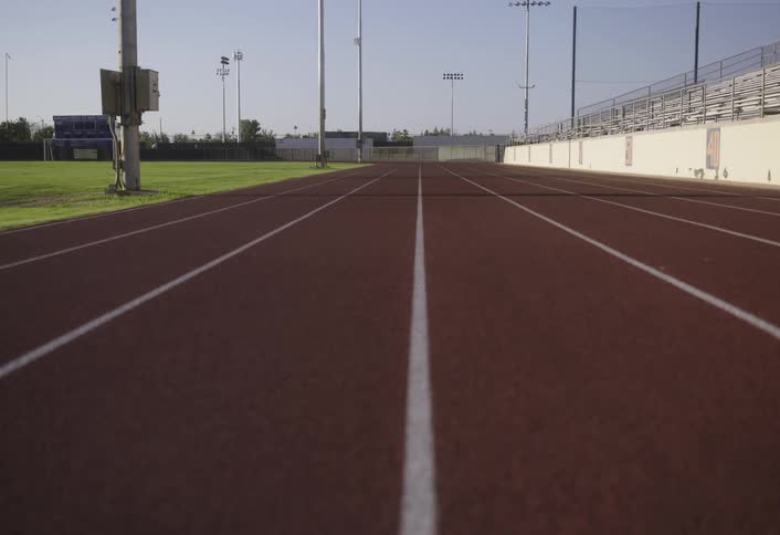 An empty running track with white lanes stretches towards the bleachers of a sunlit athletic field