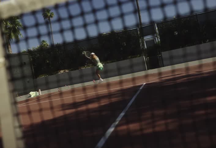 A tennis player is captured midswing on a sunny court seen through the net