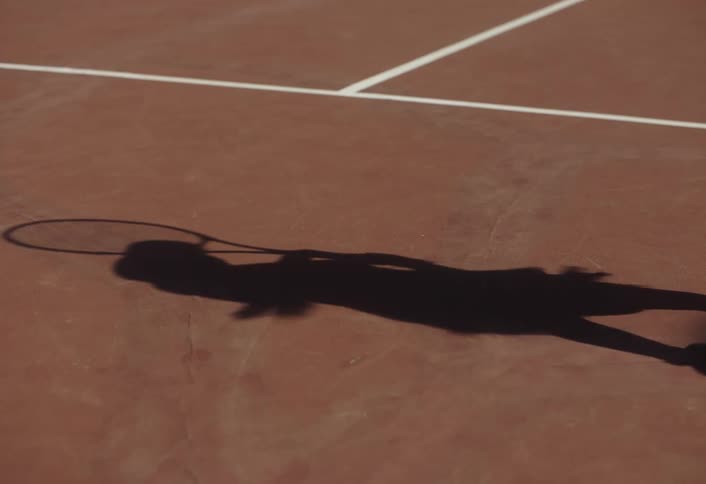 A shadow is on the court of a tennis player jumping