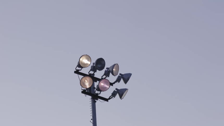 A stadium light mounted on a pole turns on and off multiple times against a cloudy sky