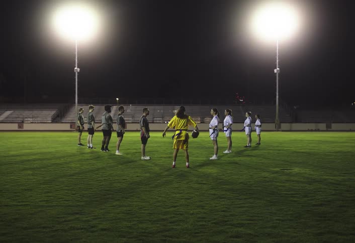 A young woman wearing a yellow jersey runs onto the field to join a group of players both men and women standing in two teams facing each other for an american football training match at night