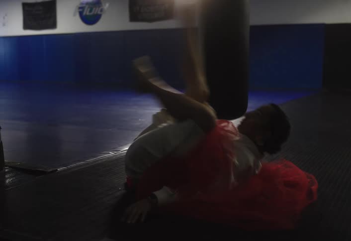 A woman in a white martial arts uniform and red tutu lies on a dark gym mat pedaling her legs in the air as part of an exercise
