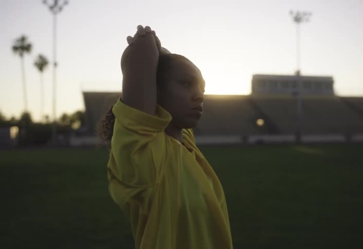 A woman in a yellow shirt is putting her arms up to stretch them and she is on a lawn of a football stadium