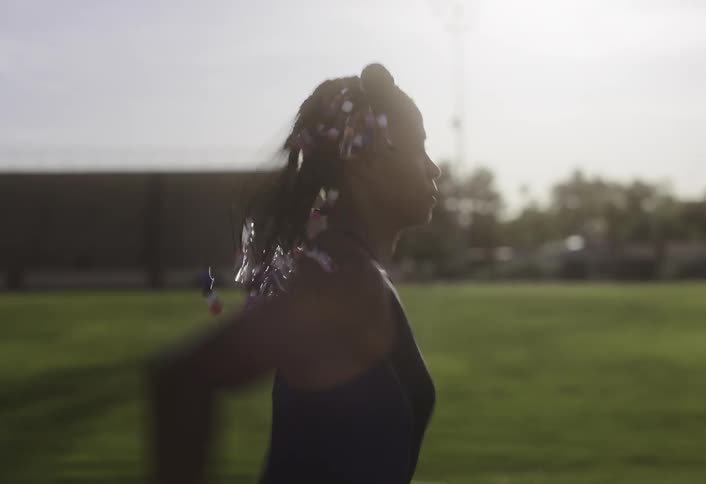 A woman with braided hair adorned with colorful beads runs across a grassy stadium under bright daylight