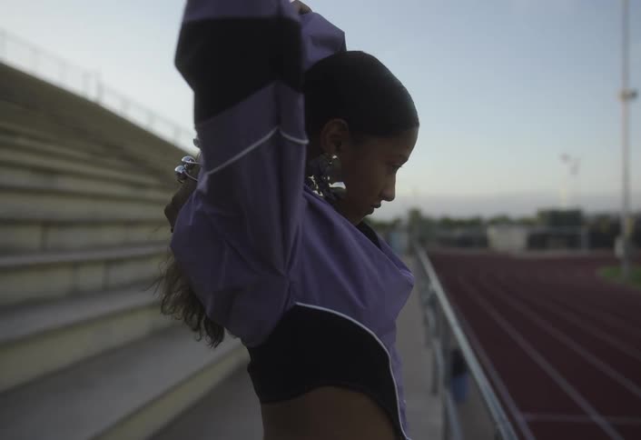 A young woman stands confidently by a stadium track looking over her shoulder at the camera