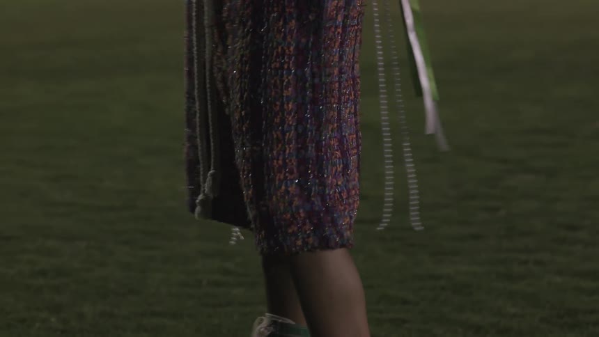 A young woman wearing green sneaker high heels a sequin skirt and a yellow football jersey with ribbons hanging from her arms stands in a mowed green football field holding a football at night