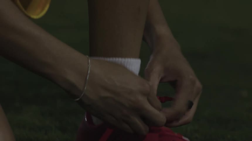 Close up of a female player tying her boot laces while kneeling on a play field at night