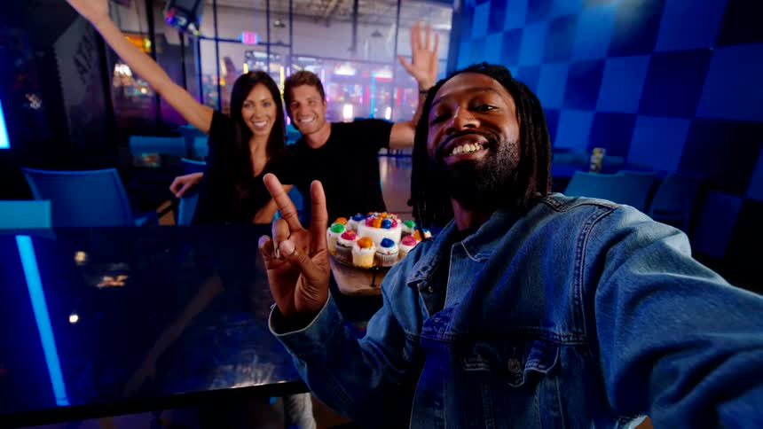 Three friends take a cheerful selfie while sitting at a table with cupcakes in a lively neonlit setting