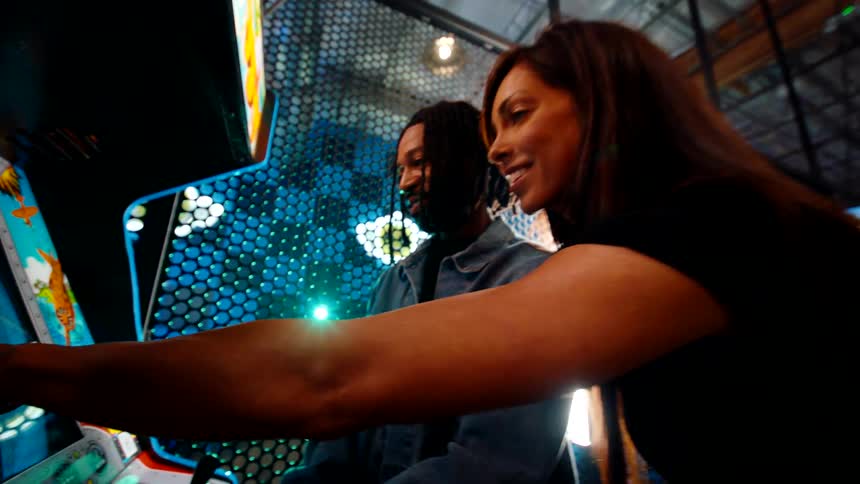 A smiling couple plays an arcade game together in a vibrant indoor setting