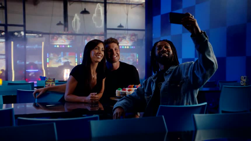 Three friends sit at a table in an arcade smiling and taking a selfie together