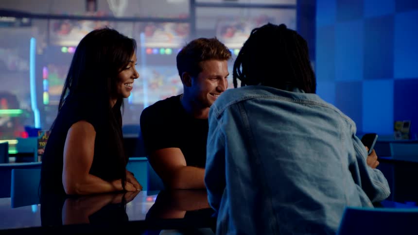 Three friends laugh and look at a phone while sitting together in a neonlit arcade bar