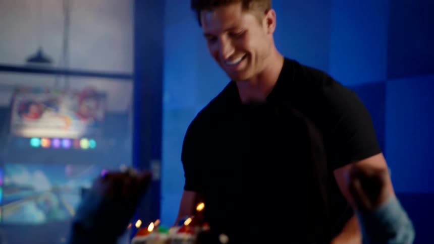 A young man brings a series of drinks to the table smiling as one of his friends cheers him on