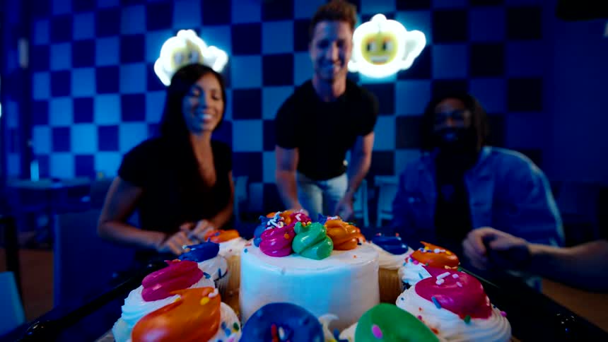 In a vibrant neon lit arcade a group of friends celebrates by grabbing colorful cupcakes from a birthday cake tray and toasting together