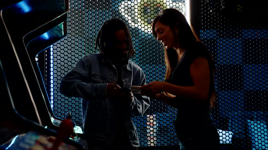 A man playing a video game at the arcade and a woman approaches to show him something on her phone