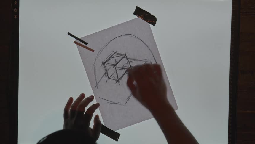 An artist sketches a geometric face outline on paper taped to a lightbox