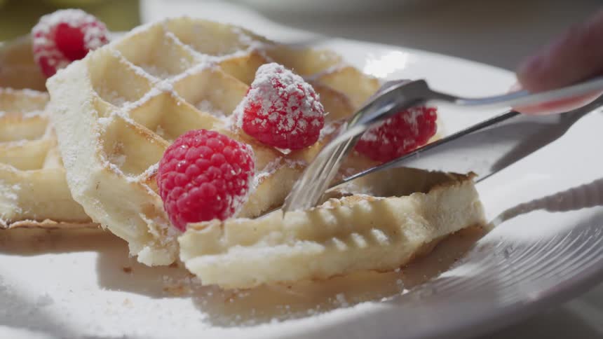 A person cuts into powdered sugardusted waffles topped with raspberries on a white plate