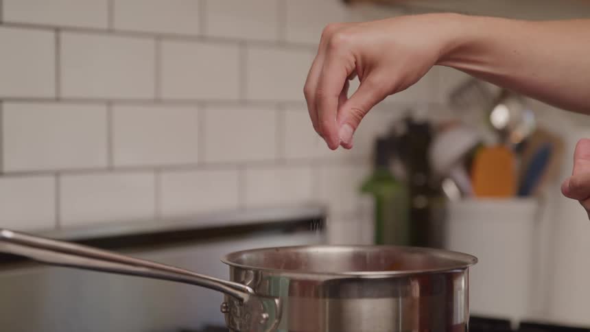 A hand sprinkles salt into a steaming pot on a kitchen stove