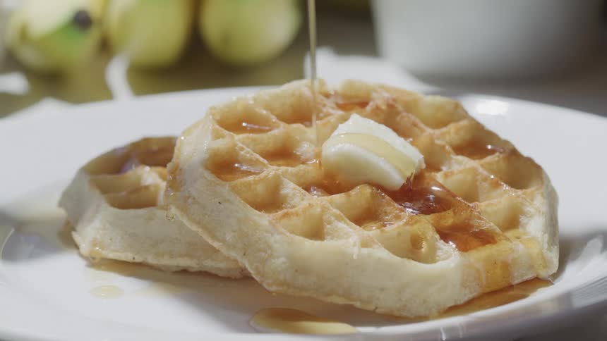 Golden syrup is being poured over two waffles topped with a pat of butter on a white plate