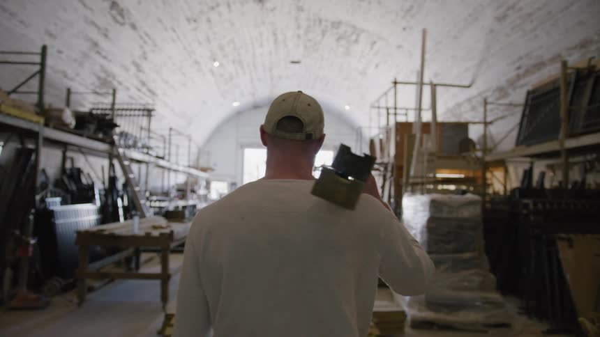 A worker walks through a warehouse carrying wooden planks on his shoulder