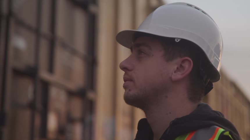 A worker wearing a white hard hat and safety vest looks at a delivery truck driving past him