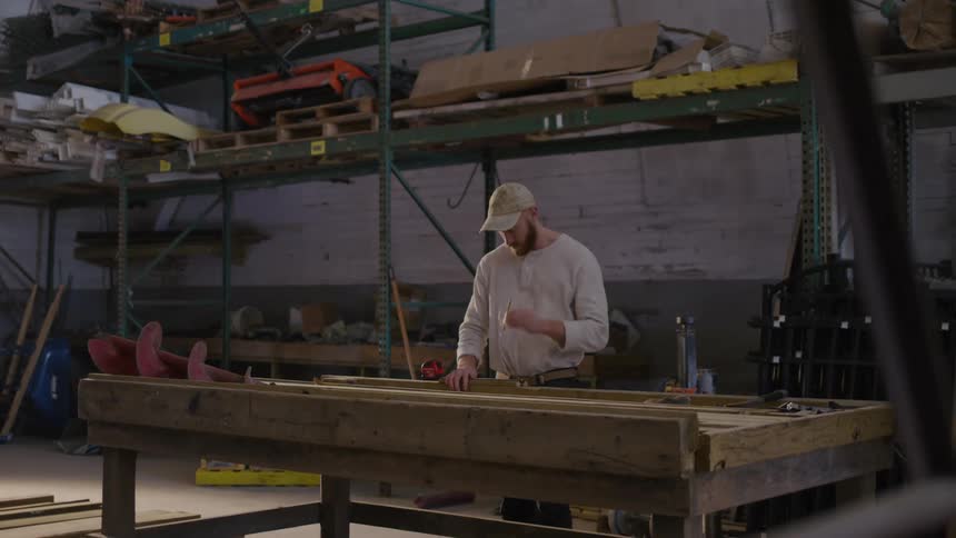 A man works on a wooden project in a warehouse workshop surrounded by tools and materials
