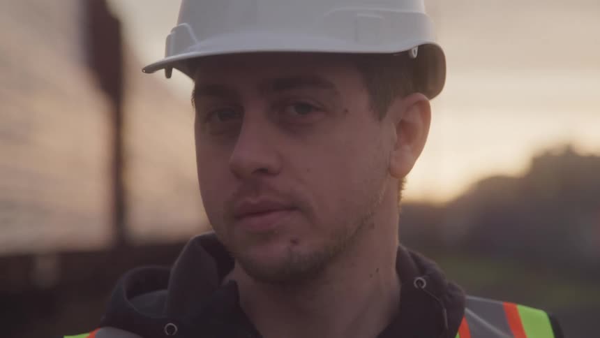 A worker wearing a white hard hat and safety vest looks at a delivery truck then turns his head away at sunset
