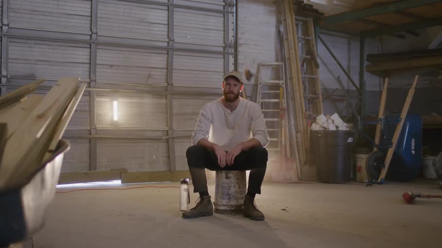 A man in black pants a longsleeve white shirt and white baseball cap is sitting on a dirty white 5 gallon bucket lost in thought in the middle of a mostly empty garage with construction equipment around him