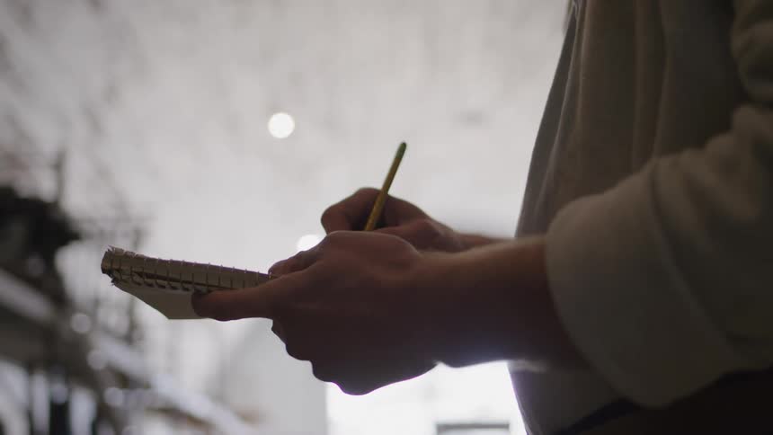 Hands of a man writing on a book while standing inside a warehouse at daytime