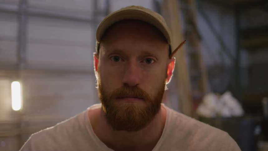 A bearded man in a cap stares at the camera inside a workshop