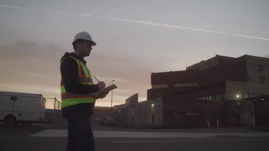 A construction worker writes on a clipboard at an industrial site during sunset