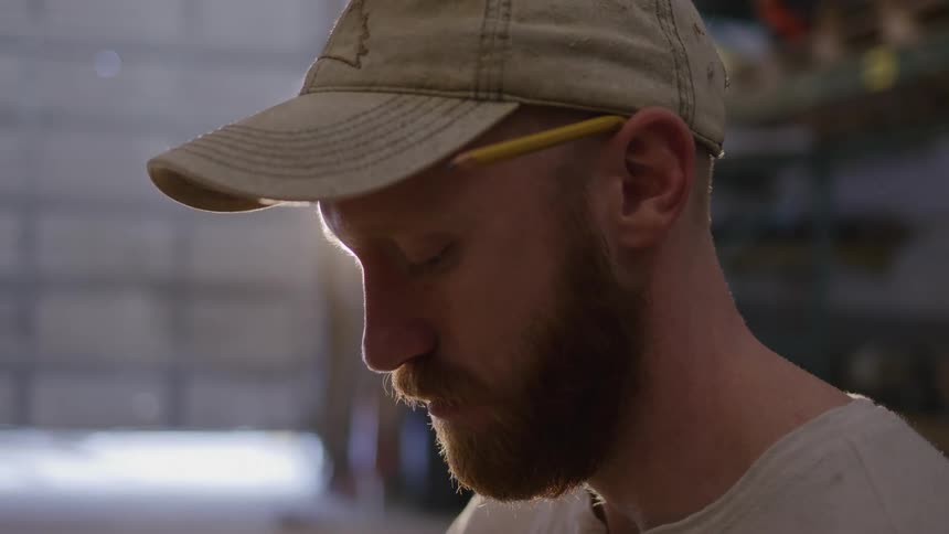 A person in a beige cap and light shirt stands indoors beside shelves stocked with assorted items taking a pencil from behind his ear
