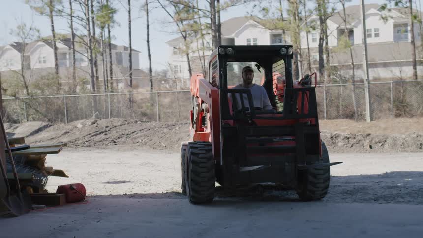 A man drives a skid steer loader out of a warehouse into a dirt lot
