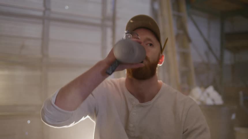 A man in work clothes drinks from a bottle inside a workshop