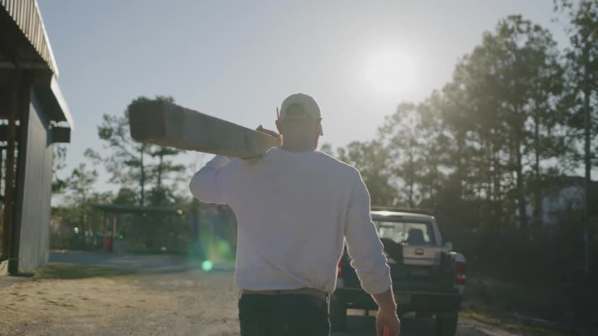 Man carries a thick wood plank on his shoulder while walking towards his truck during daytime