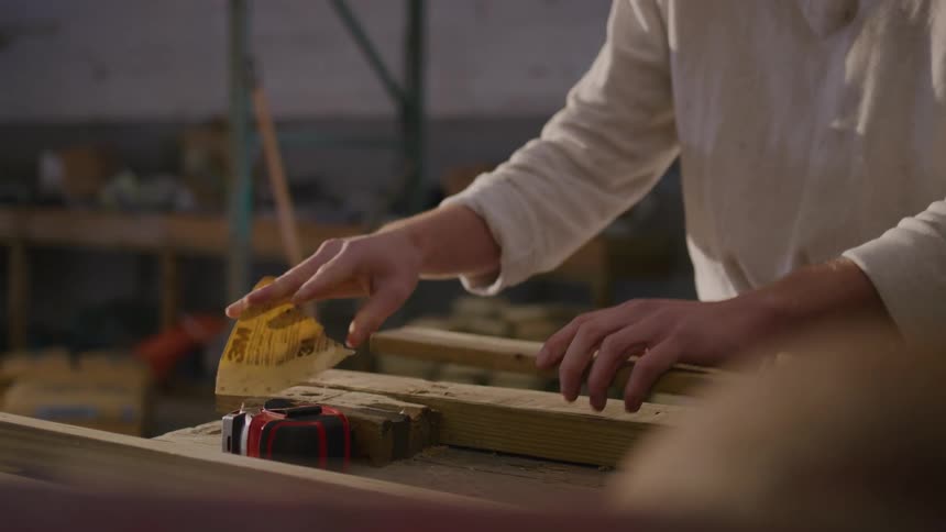A carpenter in a workshop smooths a piece of wood with sandpaper then examines it by blowing off the sawdust with a gentle breath