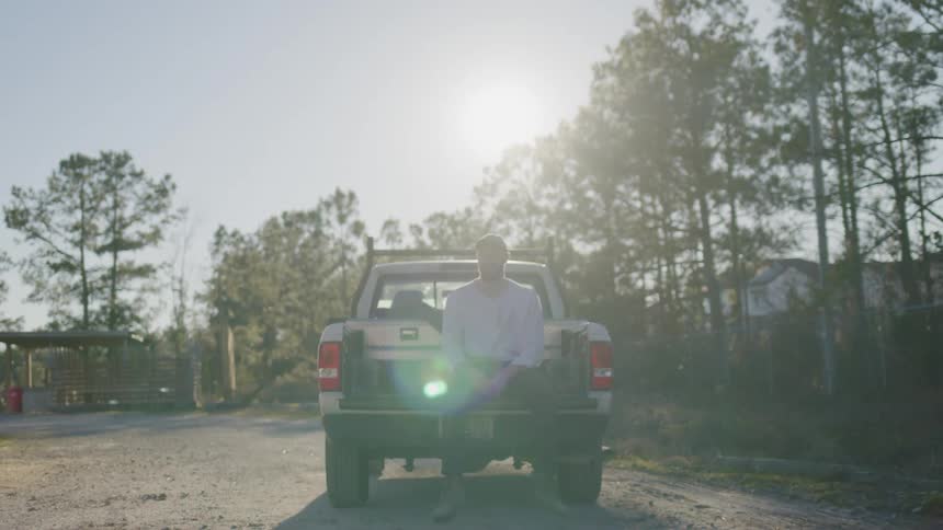 A person sits quietly on the back of a pickup truck under the bright sun
