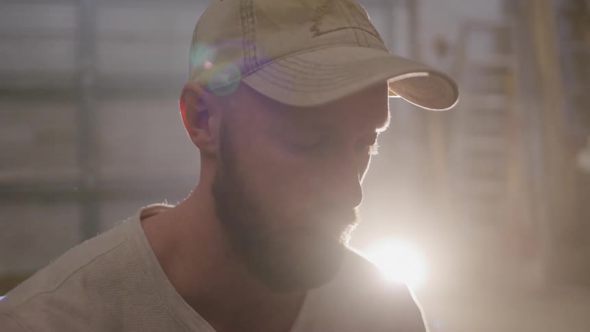The face of a man with a beard and wearing a cap and he is looking at something that is below him in an indoor environment and a natural light is coming from behind