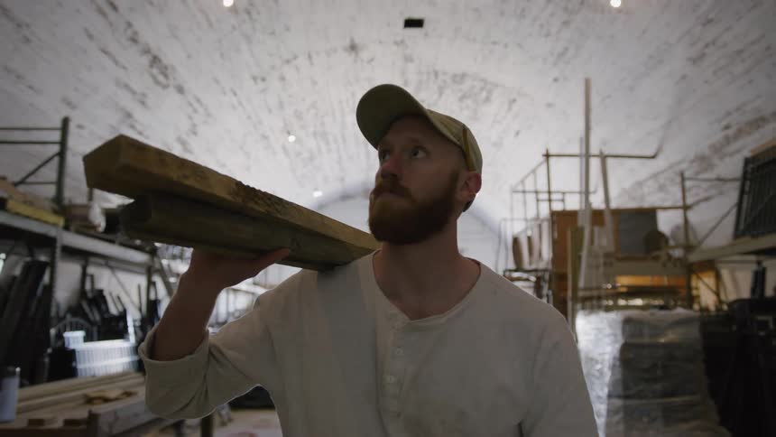 Close up of a male carpenter carrying two wood planks over his shoulder while walking inside a workshop