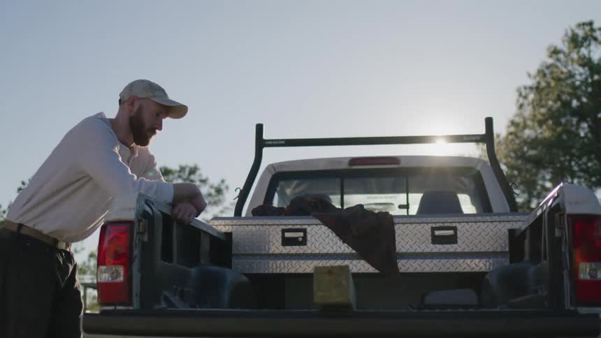 A man leans on the tailgate of a pickup truck under the sun