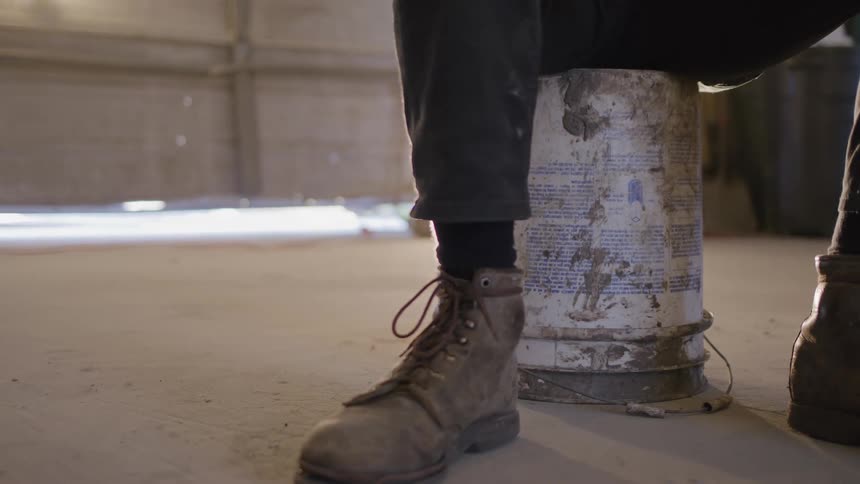 A person wearing green boots sits on a bucket in a garage drinking from a water bottle