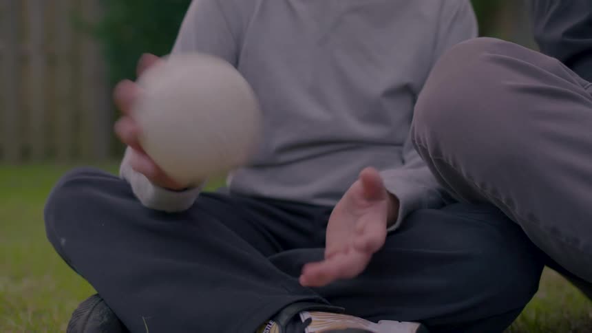 A person sits on a lawn at midday bouncing a white ball while sitting next to another person