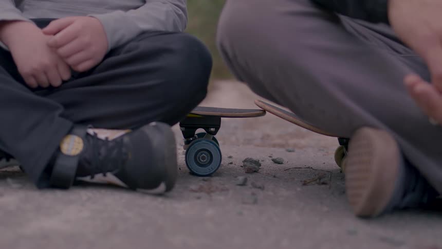 Two boys wearing longsleeved shirts skinny pants and sneakers are sitting crosslegged on skateboards one much younger than the other rubbing their hands