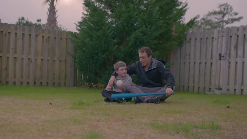 Father and son sit on the grass in a fenced backyard holding a blue baseball bat and a ball