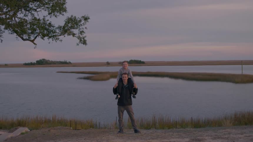 Father carrying son on shoulders posing for a photo on lake shoreline at dusk