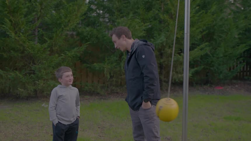 Young man converses with a boy as they both stand on a lawn outdoors during daytime
