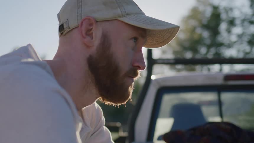 Man on face cap and white shirt rests on the back of a parked truck at daytime