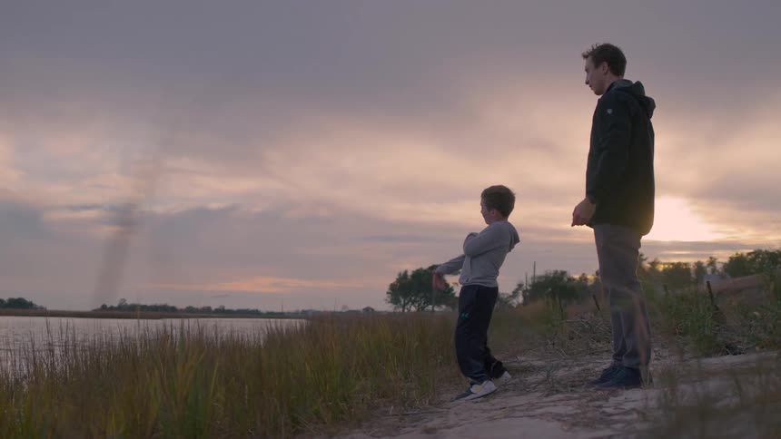 Boy playing stone skipping while standing with a young man at the river bank at sunset
