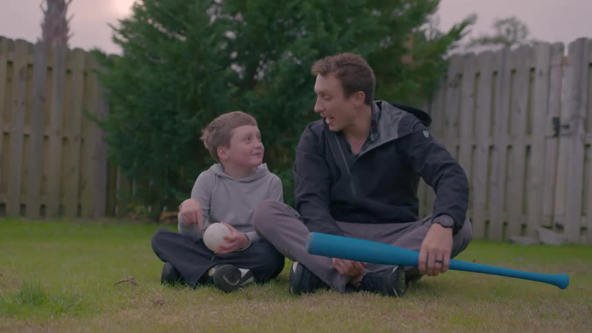 A father and son sit on the grass having a conversation while holding a ball and bat