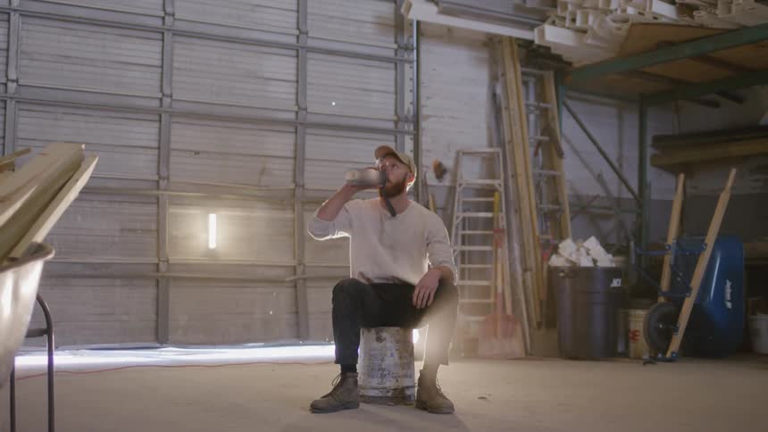 A man sits on a bucket in a workshop drinking from a bottle under soft industrial lighting