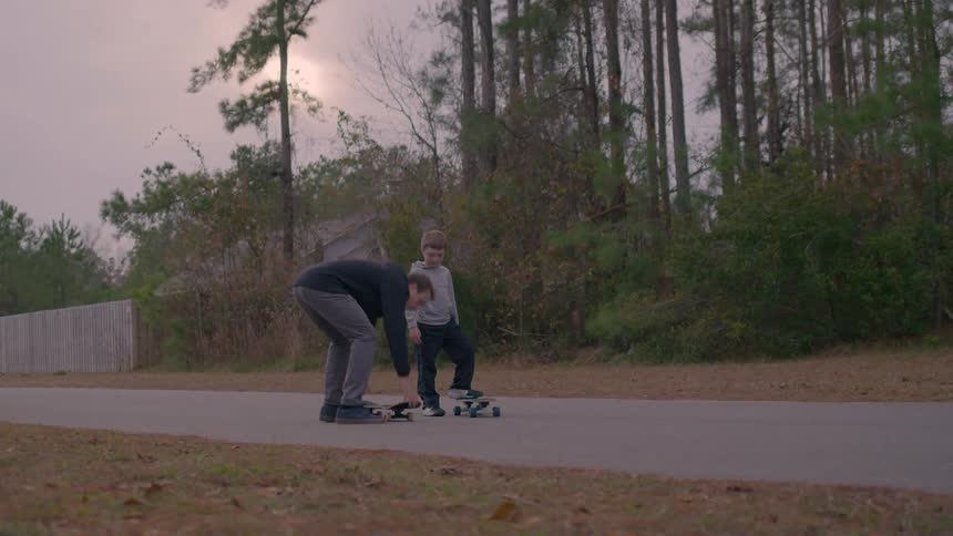 A man wearing a black jacket teaches a kid how to ride a skateboard on the sidewalk at sundown